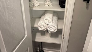 Um interior claro com uma porta, uma prateleira com toalhas enroladas e um objeto preto na prateleira.