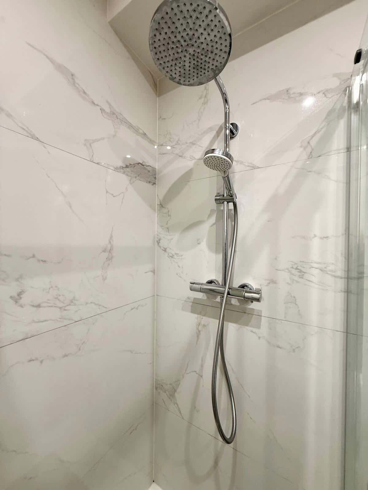 Eine moderne Dusche mit Handbrause und fest installierter Duschleiste, vor einer Marmorwand.