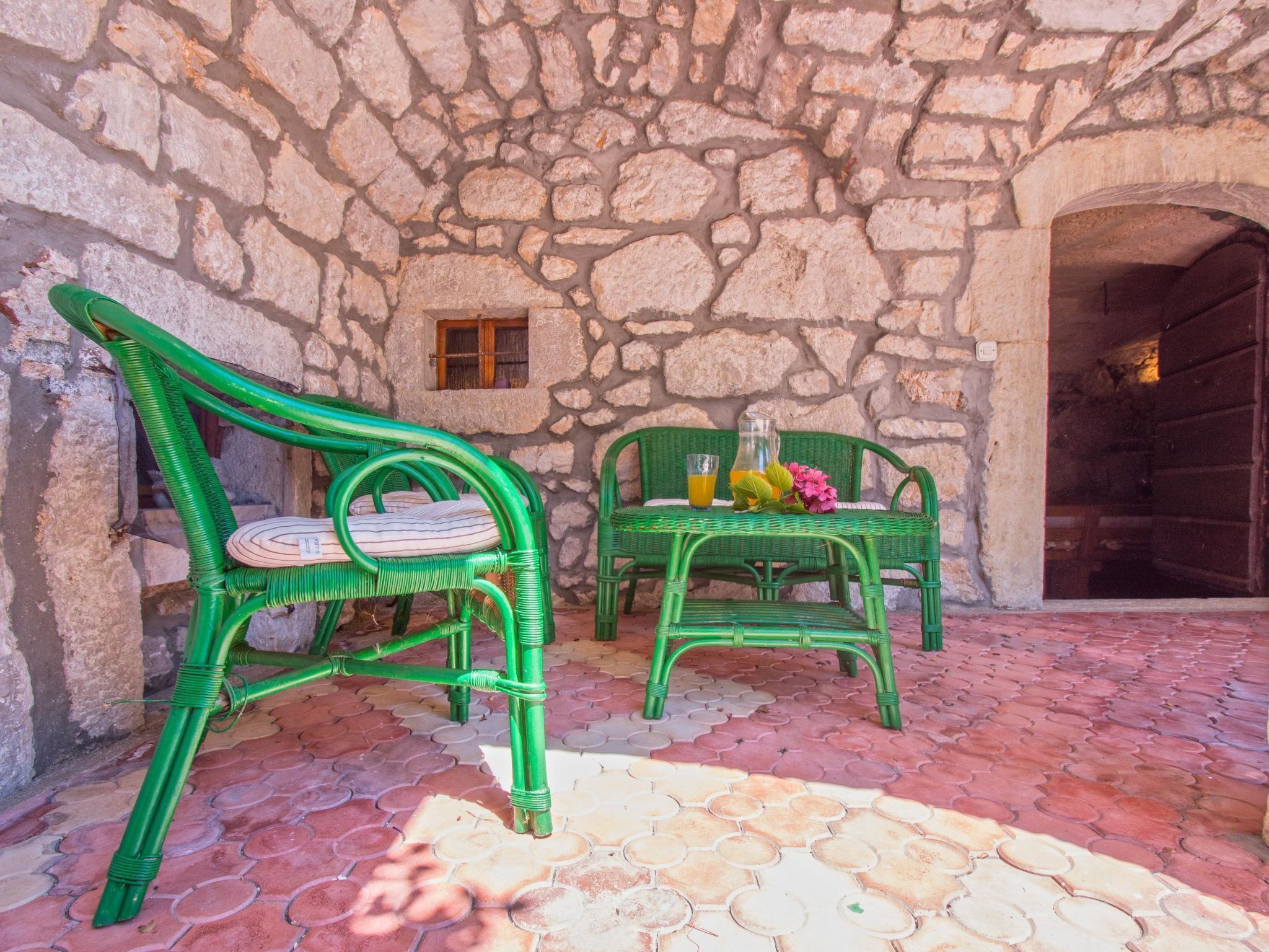 A stone wall exterior with two green wicker chairs and a table, a small window, and a door.