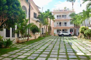 A paved driveway leading to a multi-story building with balconies, surrounded by lush greenery and palm trees. - Steps from the Beach · 1BR w/ Rooftop Jacuzzi (Punta Cana)