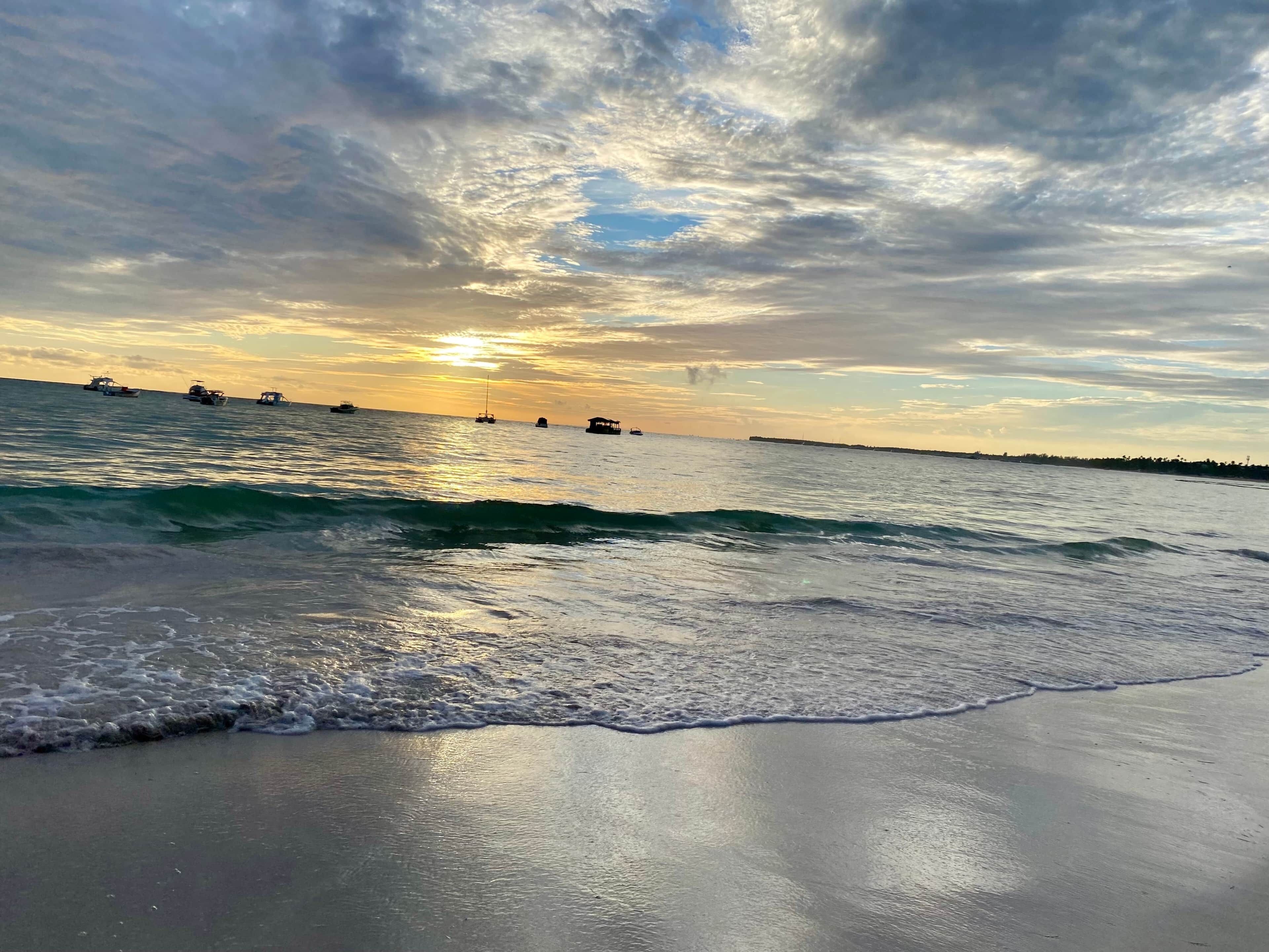 A beach with waves and boats in the distance under a cloudy sky.