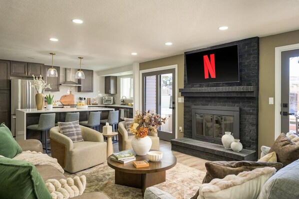 Ein modernes Wohnzimmer mit Kamin, einem an der Wand befestigten Fernseher und einer Küche im Hintergrund.