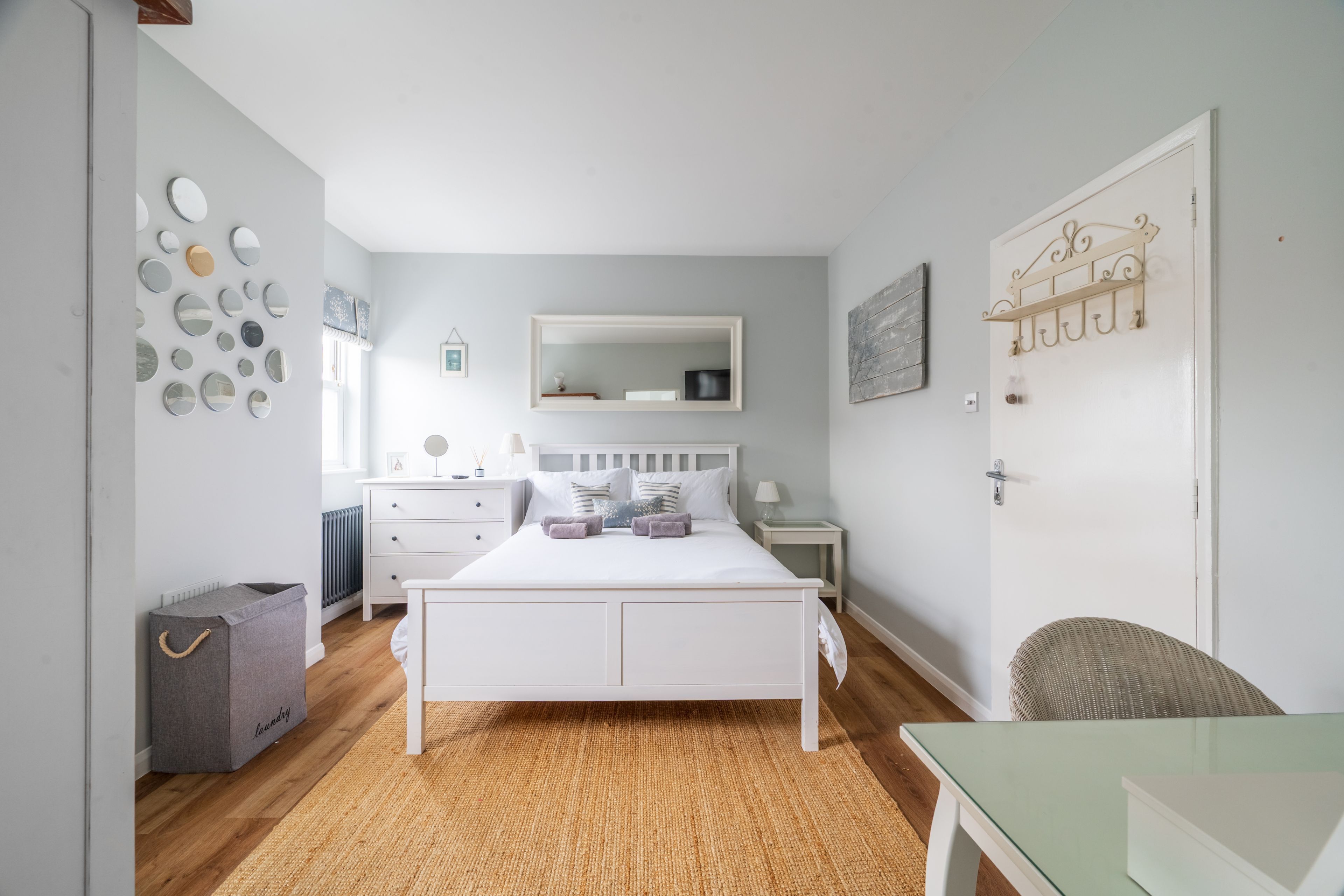 Un dormitorio con cama, cómoda, mesita de noche, silla, mesa, radiador, cesto de ropa y un muro con espejos y un cuadro.