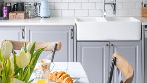 Ett kök med en vit bänkskiva, en diskho och skåp. Där finns en trästol med ett ryggstöd i metall, ett bord med en vit duk, ett glas apelsinjuice, en croissant och en mugg med ett motiv av en cupcake.