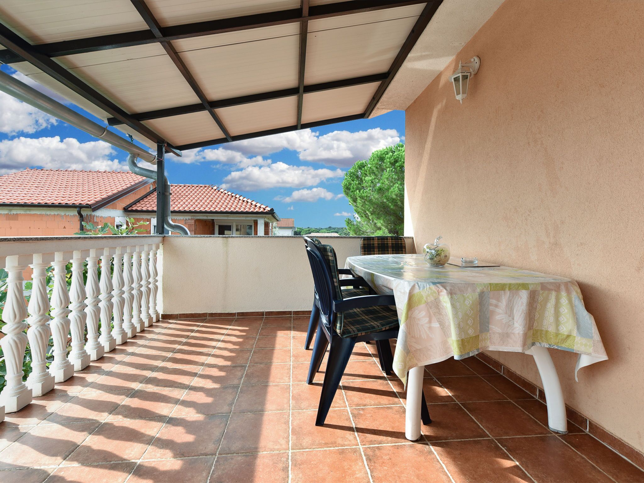A covered patio with a table and chairs, a white balustrade, and a view of houses with red-tiled roofs.