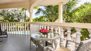 Un balcón con muebles de mimbre, una mesa de vidrio con un centro de mesa floral y exuberante follaje tropical.