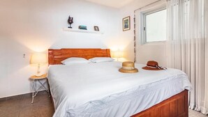 Una cama bien tendida con sábanas blancas, cabecera de madera y un sombrero de paja sobre la cama. Hay una mesita de noche con una lámpara y una ventana con cortinas.
