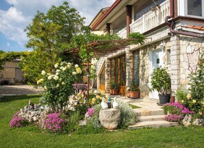 Un bâtiment en pierre doté d’un balcon, entouré d’un jardin bien entretenu où poussent diverses plantes et fleurs.
