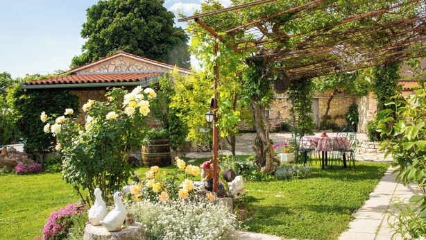 Um jardim com uma pérgola, um edifício de pedra e uma mesa com cadeiras.