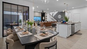 A modern kitchen with a central island, a dining area with a long table set for six, and a living area with a sofa and TV. - 9096 Amazing house 6/5 W/Pool Solara Resort (Kissimmee)
