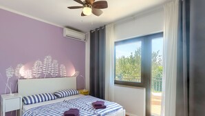 A bedroom with a bed, a ceiling fan, an air conditioning unit, a nightstand, a lamp, and a window with curtains. - Villa Valbandon by Interhome (Fažana)