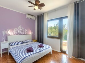 Ein Schlafzimmer mit Bett, Deckenventilator, Klimaanlage, Nachttisch, Lampe und einem Fenster mit Vorhängen.