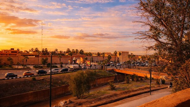 Un paisaje urbano con un muro destacado y palmeras, una calle concurrida con coches, y un cielo al atardecer.