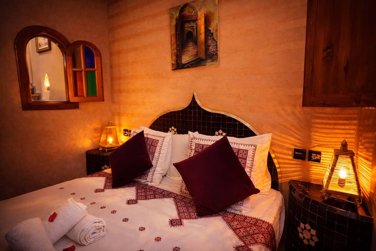 A hotel room with a bed, pillows, a nightstand, a mirror, a wall painting, and a tiled wall.