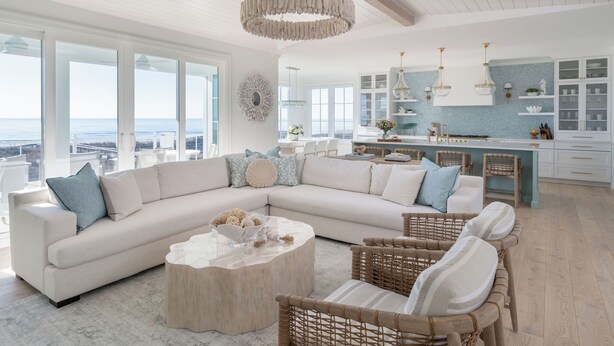 A spacious living room with a large L-shaped sofa, a round coffee table, and wicker chairs. A kitchen with white cabinets and blue backsplash is visible in the background.