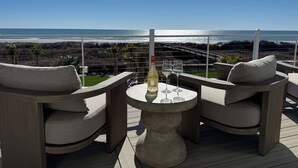 A balcony with two wicker chairs, a small round table, and a bottle of wine. The view includes a beach and ocean.