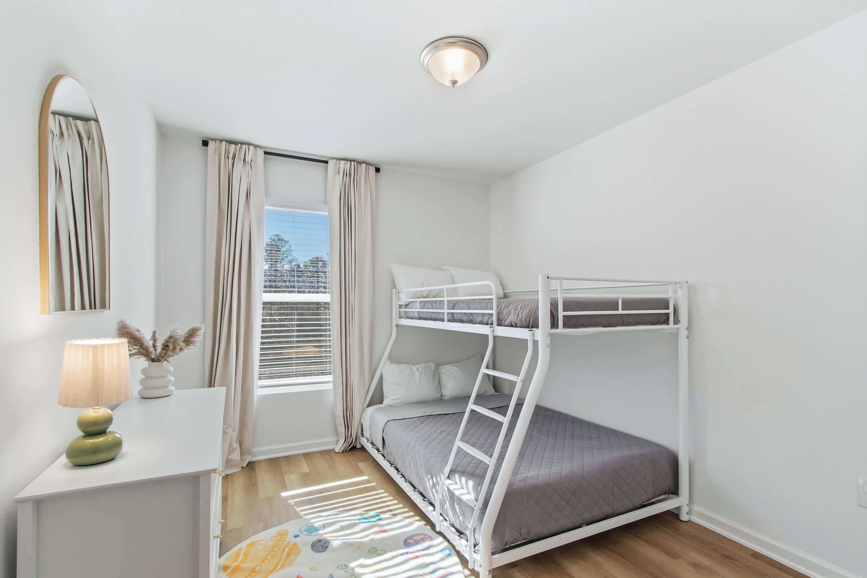 A bunk bed room with a desk, lamp, and window with curtains.