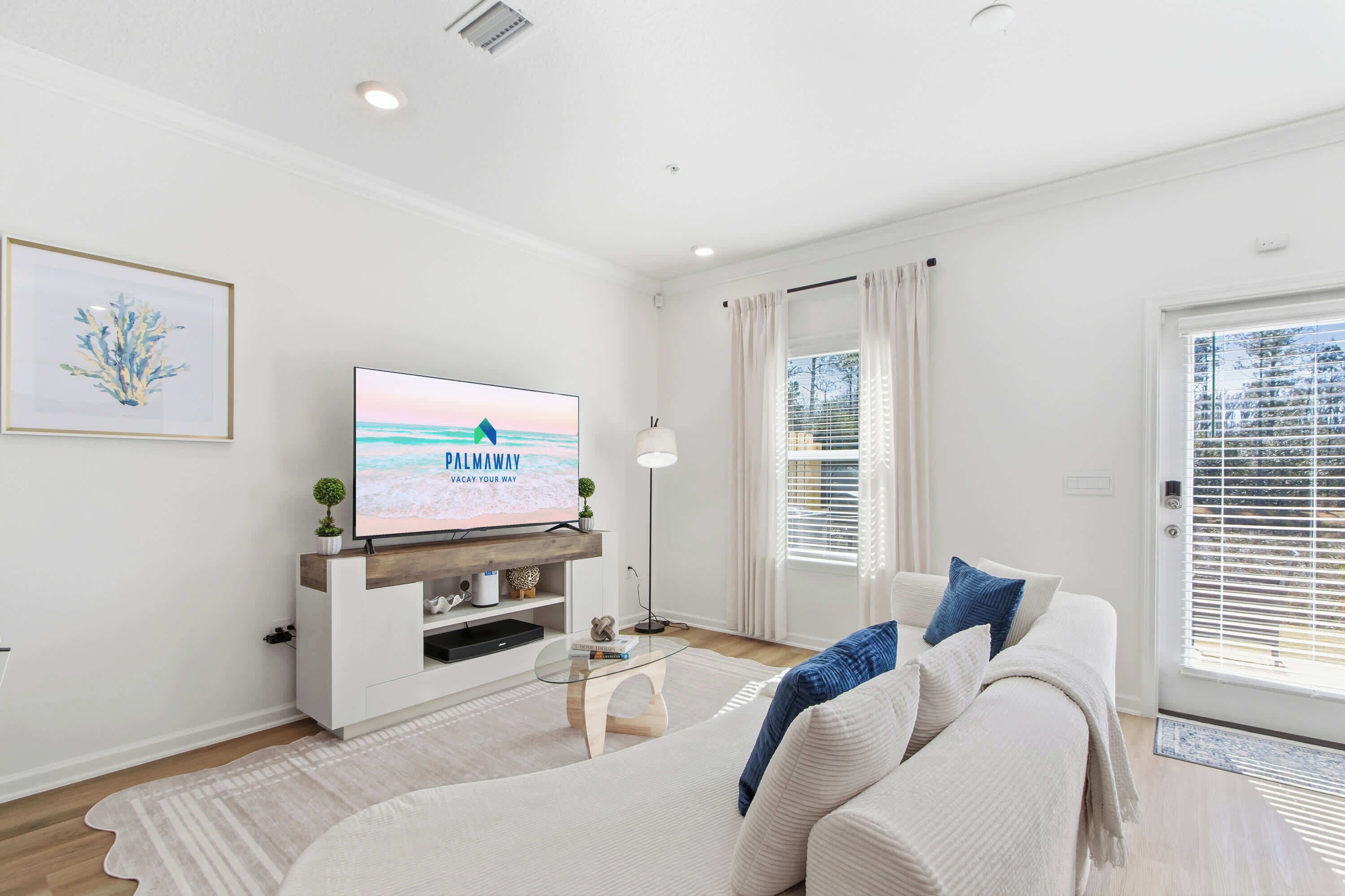 A modern living room with a flat-screen TV, a sofa, a coffee table, and a large window with blinds.