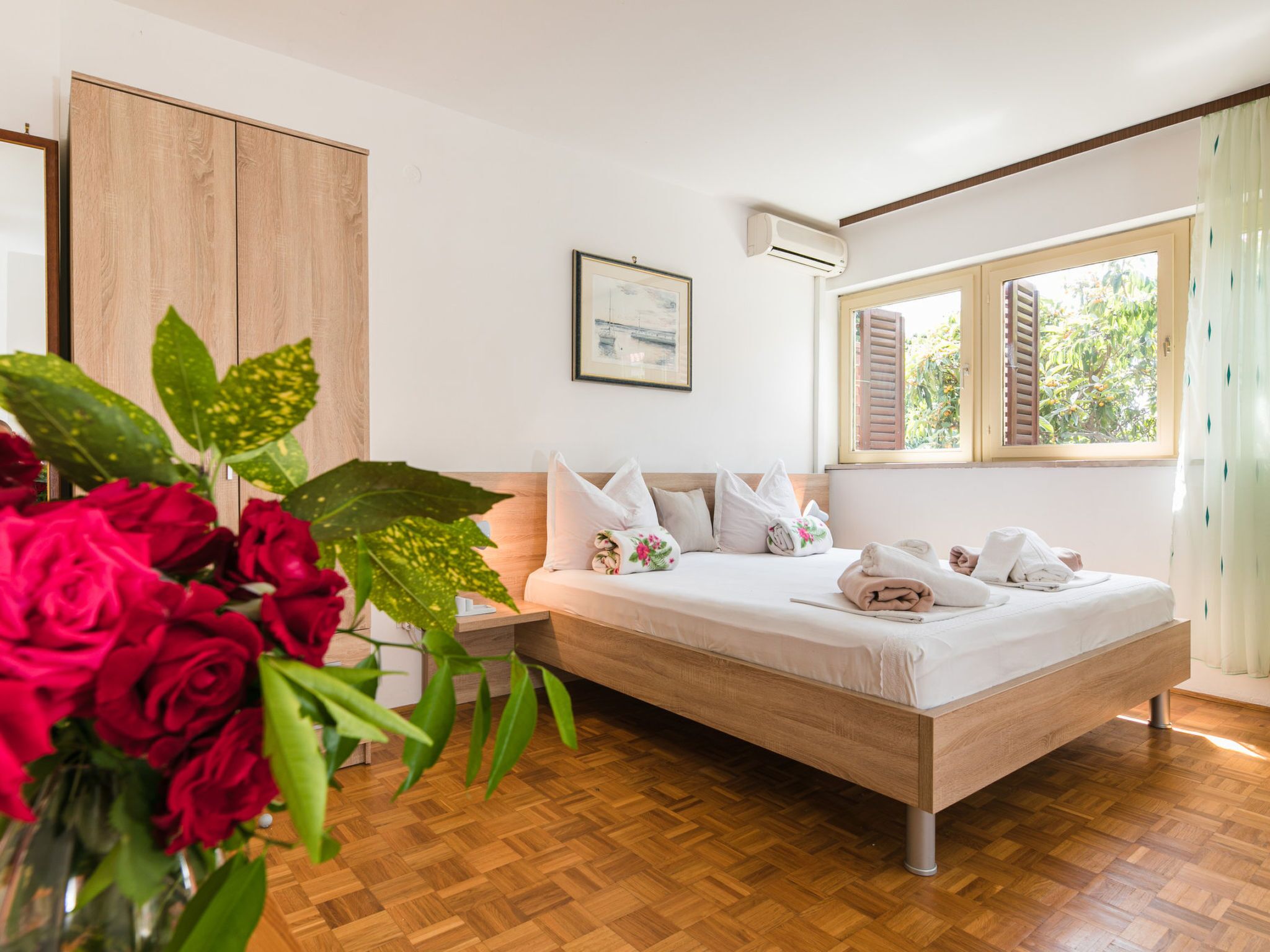 A bedroom with a wooden bed, white bedding, a bedside table, a framed picture, and a window with shutters.