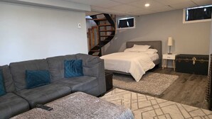 A modern bedroom with a grey sofa, a bed with white bedding, a small bedside table with a lamp, a suitcase, and a TV mounted on the wall. - Charming Waterfront Cottage –  Fully Equipped Kitchen & Fast Wi-Fi (Plaisance)
