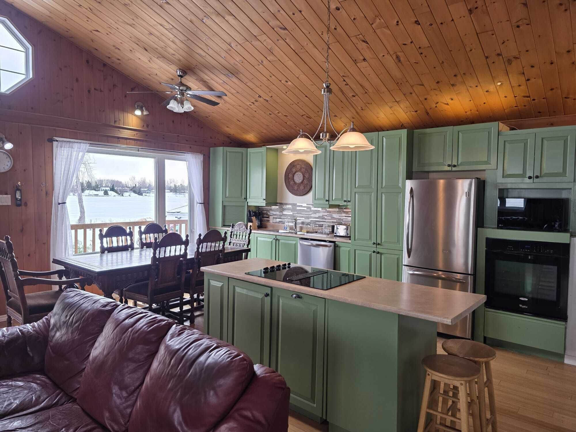 A cozy cabin-style kitchen with green cabinets, a stainless steel refrigerator, and a dining area with a wooden table and chairs. - Charming Waterfront Cottage –  Fully Equipped Kitchen & Fast Wi-Fi (Plaisance)