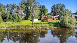 A lakeside property with a house, a small shed, and dense surrounding trees. - Charming Waterfront Cottage –  Fully Equipped Kitchen & Fast Wi-Fi (Plaisance)