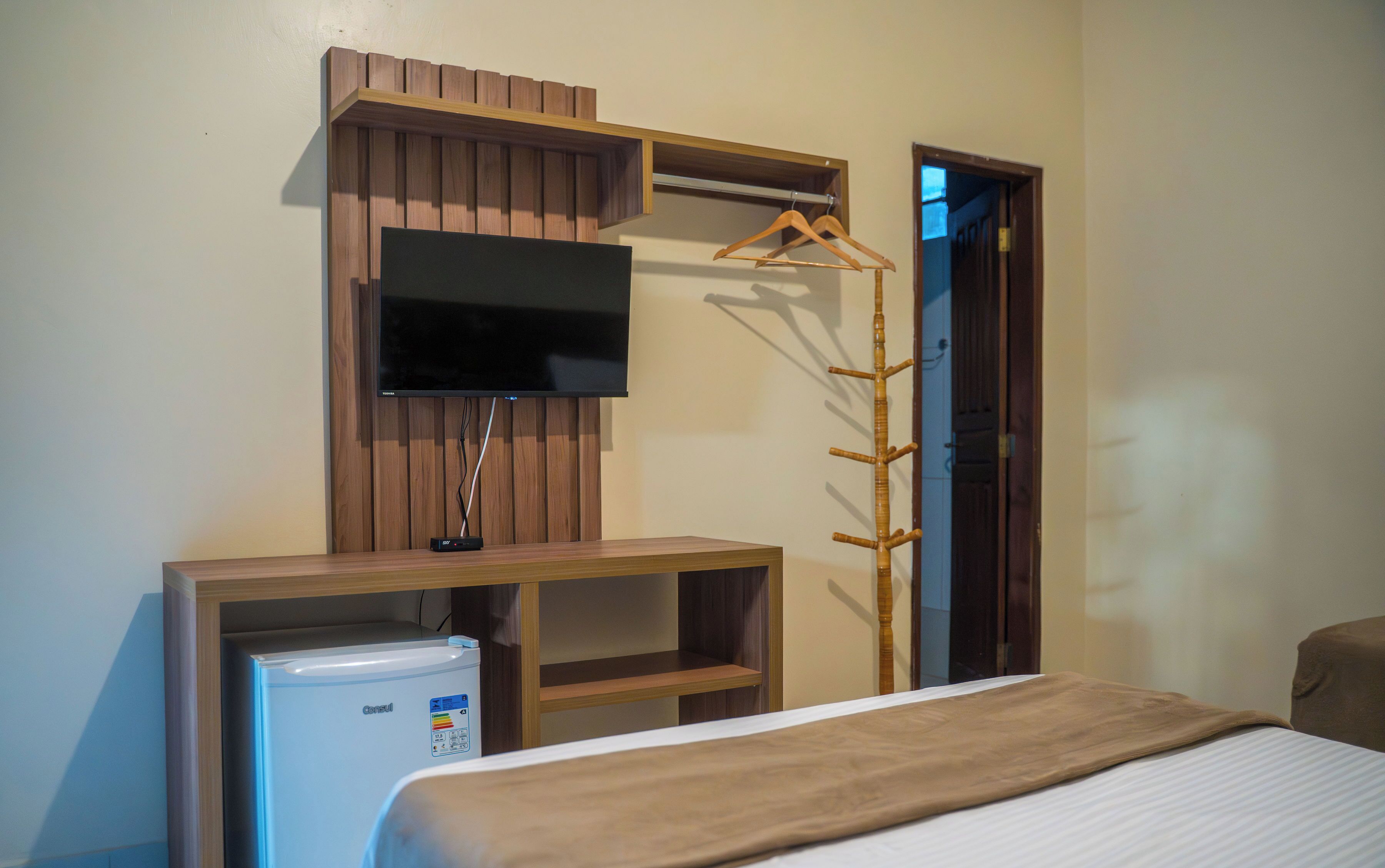 A hotel room with a bed, a TV mounted on a wooden panel, a small fridge, and a coat rack.
