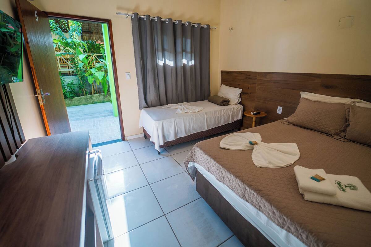 A hotel room with two beds, a wooden headboard, a small bedside table, and a view of a garden through an open door.