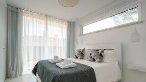 Quarto moderno com uma cama grande, roupa de cama branca e vista para o exterior através de cortinas leves.