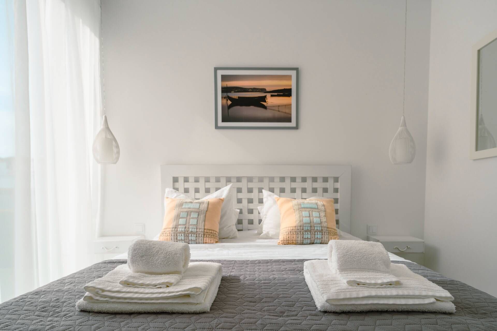 Een keurig opgemaakt bed met een grijze deken, twee witte handdoeken en een lijst met een foto van een zonsondergang boven het water aan de muur.