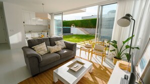 Uma sala de estar moderna com um sofá cinzento, mesa de centro branca e vista para um jardim verde através de grandes portas de correr de vidro.