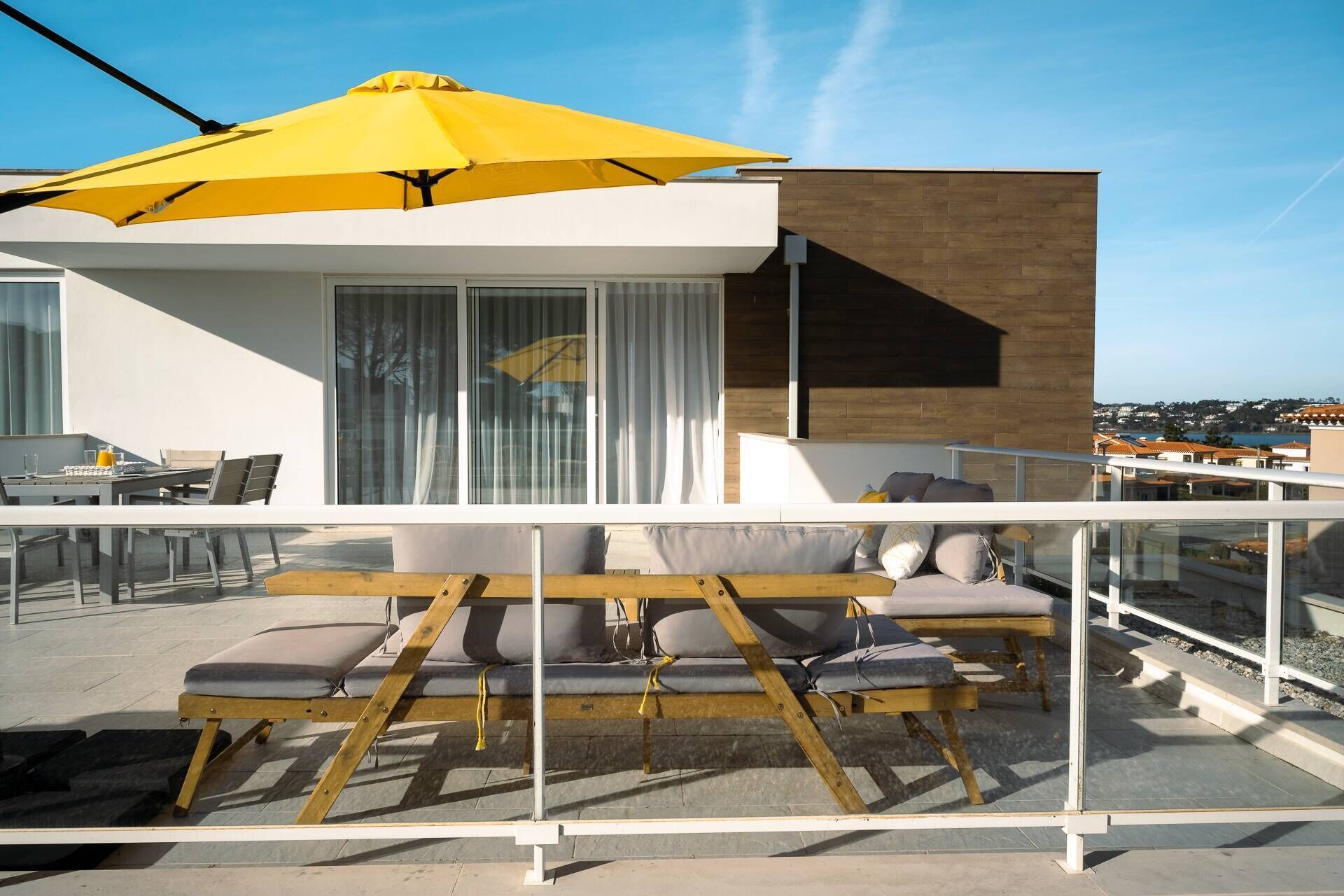 A modern outdoor seating area with a yellow umbrella, a wooden bench, and a sofa with cushions.
