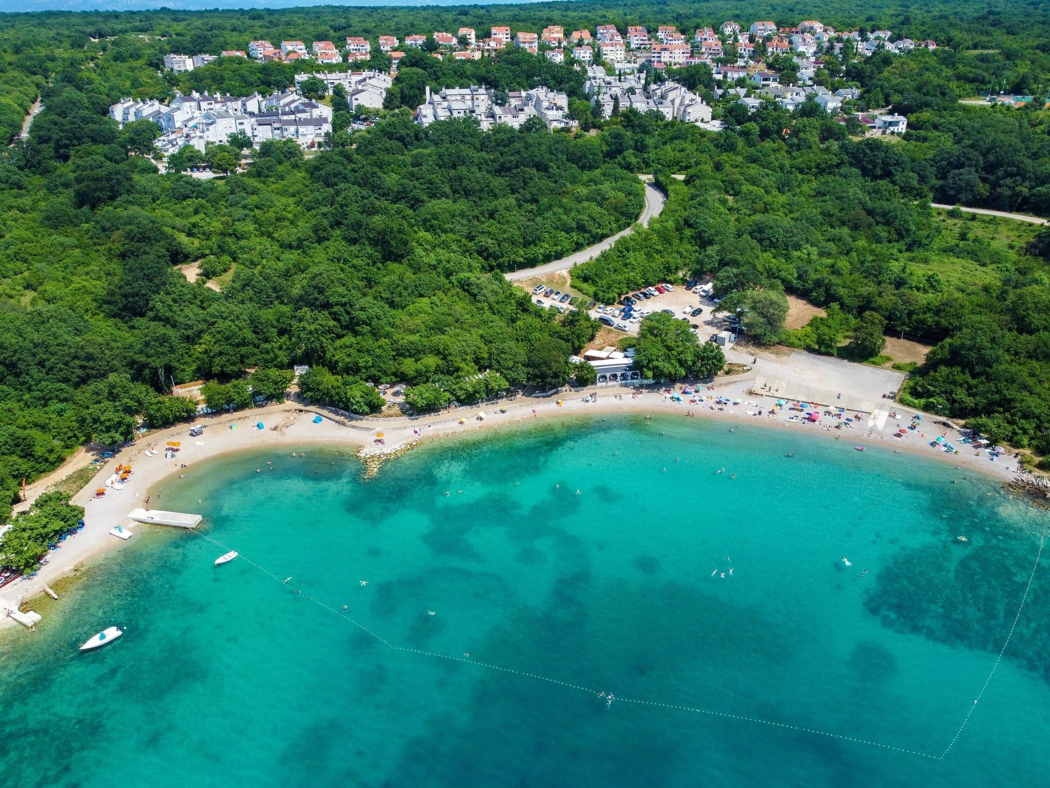 Vista aérea de una zona costera con playa, aguas turquesas cristalinas y una área residencial con casas y vegetación al fondo.