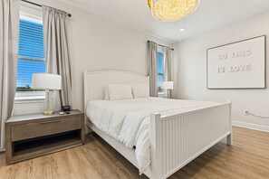 Une chambre à coucher avec un lit blanc, des tables de chevet, une lampe fixée au mur et une fenêtre avec des rideaux.