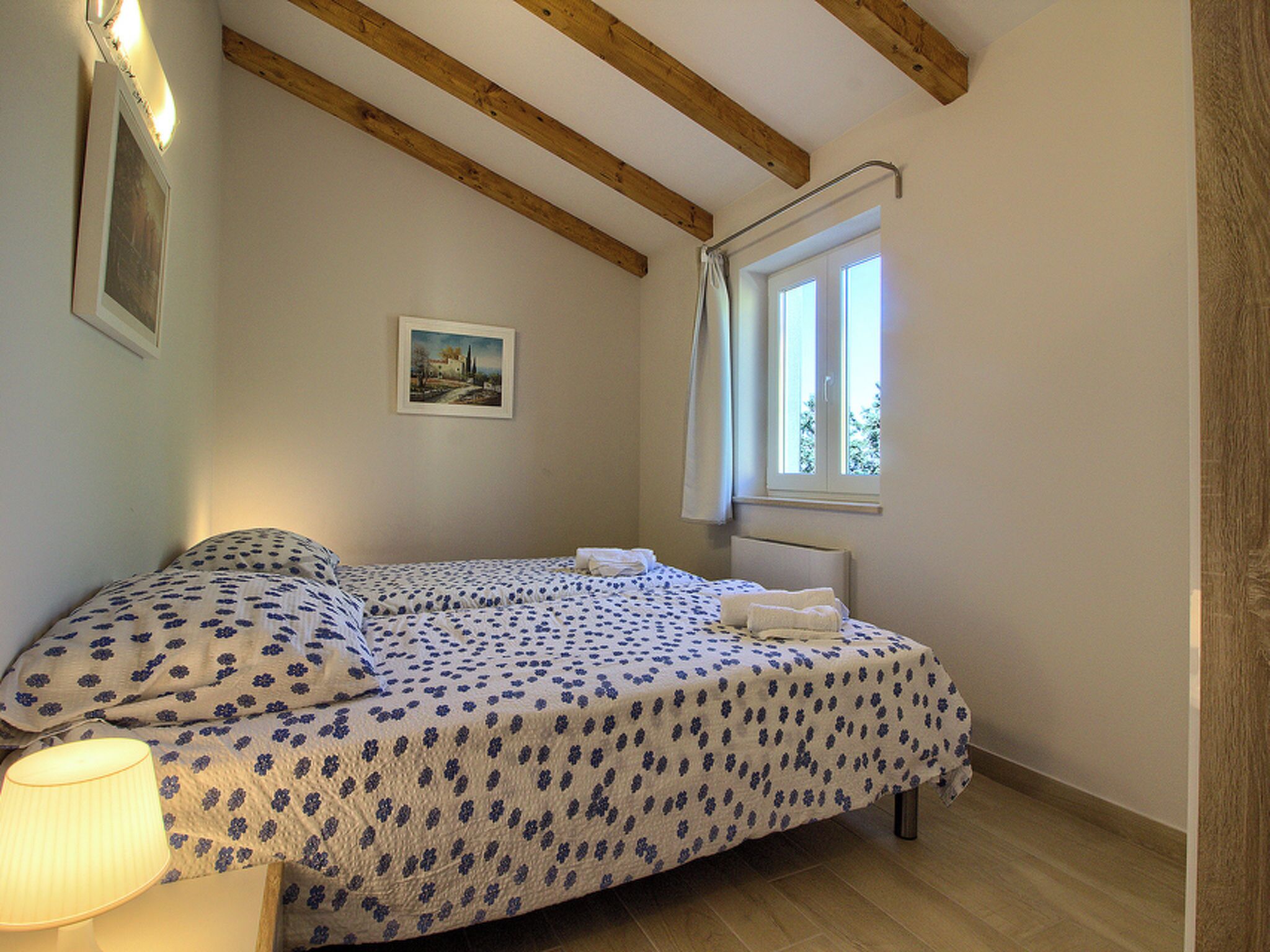 Un dormitorio con cama, ventana, un cuadro y vigas de madera en el techo.