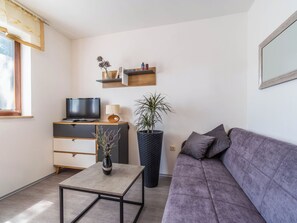 Ein modernes Wohnzimmer mit grauem Sofa, Couchtisch, Fernseher, Bücherregal und einer großen Topfpflanze.