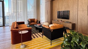 Ein modernes Wohnzimmer mit Sofa, Sessel und Couchtisch, einem Fernseher an der Wand und großen Fenstern mit Vorhängen.