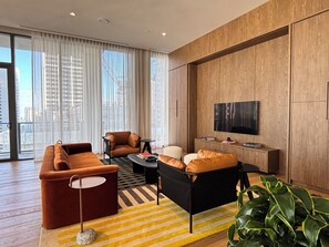 Uma sala de estar moderna com sofá, poltrona e mesa de centro, uma TV na parede e janelas amplas com cortinas.