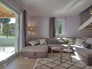 Un salon moderne avec un canapé, un pouf et une table basse, une lampe, et une vue sur l’extérieur.