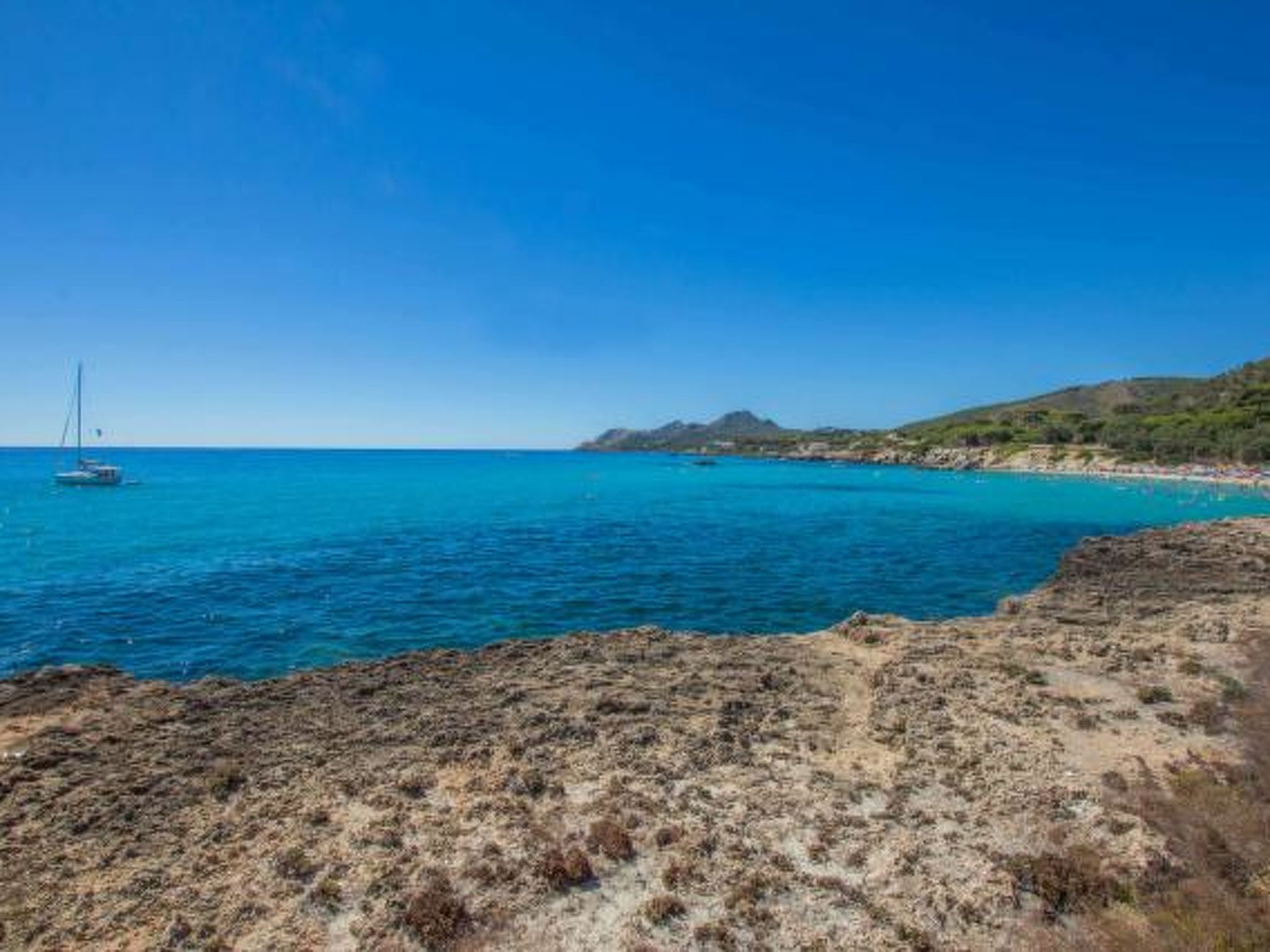 A coastal landscape with a clear blue sea, rocky terrain, and a distant mountain.