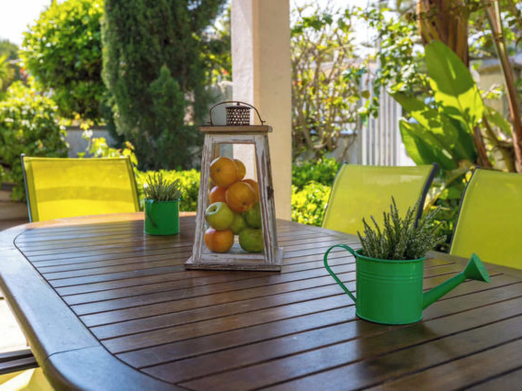 Una mesa de madera con un farol de frutas, macetas verdes y sillas amarillas.