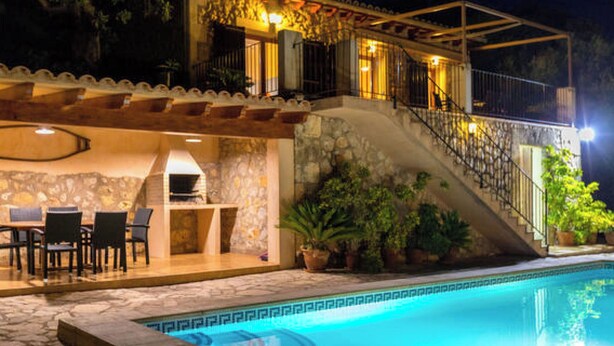Uma área à beira da piscina com um muro de pedra, assentos externos e uma escadaria que leva a uma varanda.