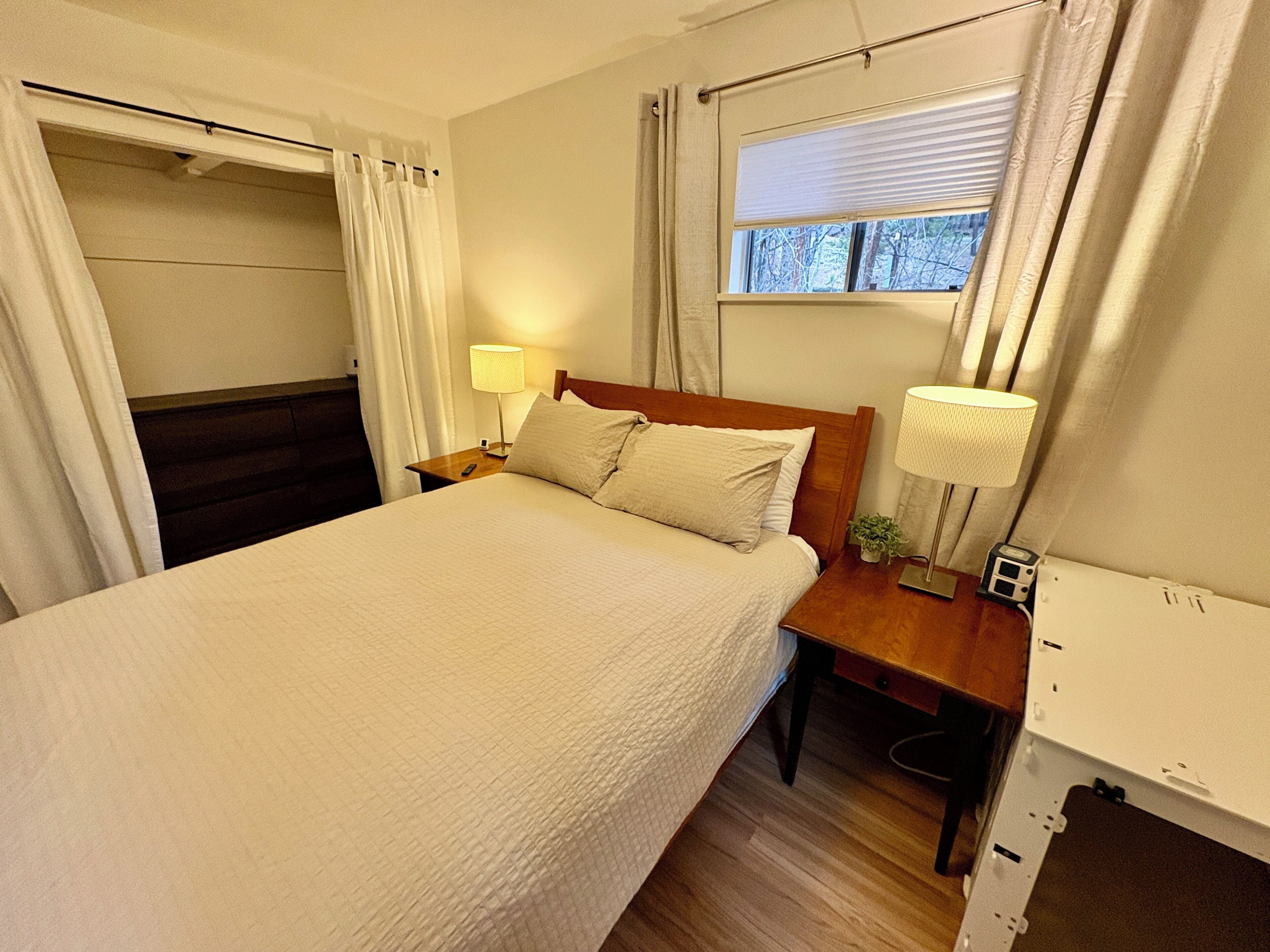 Una cama bien hecha con tablero de madera, una mesita de noche con lámpara y una ventana con cortinas.