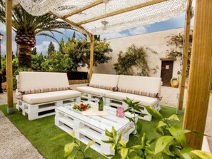 Ein überdachter Außenbereich mit weißen Sofas und einem Couchtisch, umgeben von Grünpflanzen und blauem Himmel.