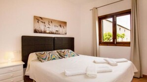 A neatly made bed with white linens, a headboard, a bedside table with a lamp, and a window with curtains. - Manyo Petit by Interhome (Sa Pobla)