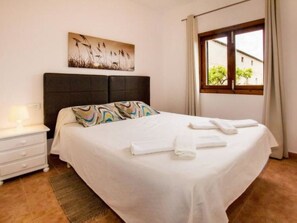 Ein ordentlich bezogenes Bett mit weißen Leinen, ein Kopfteil, ein Nachttisch mit Lampe und ein Fenster mit Vorhängen.