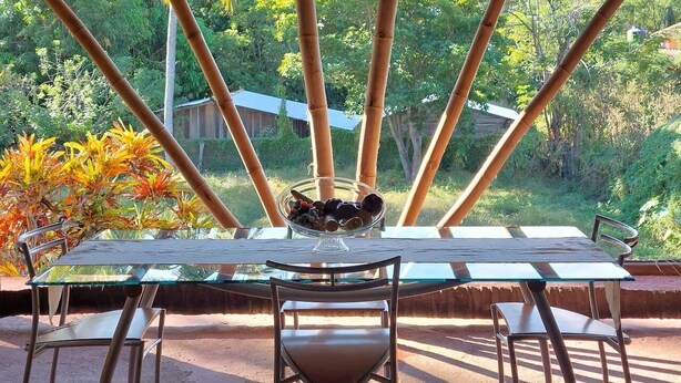 Een buiten eethoek met een tafel en stoelen, een fruitschaal, en uitzicht op een weelderige tuin en bomen.
