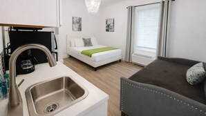 Ein modernes Hotelzimmer mit Küchenzeile, einem Bett mit grüner Tagesdecke, einem grauen Sofa und einem großen Fenster mit Vorhängen.