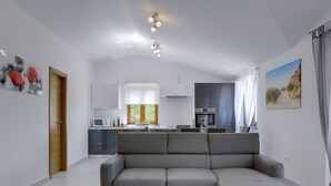 Un salon moderne avec un canapé d’angle gris, une cuisine avec des meubles blancs et un coin repas comprenant une table et des chaises.