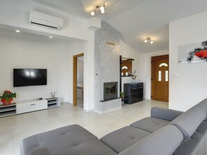 Ein modernes Wohnzimmer mit Flachbildfernseher, grauen Sofas, Kamin und einem zur Schlafzimmer führenden Gang.